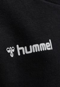 Tessuto nero con una trama texturizzata, caratterizzato da un logo "hummel" bianco e un'emblema a forma di ape, posizionato vicino all'angolo in basso a destra.