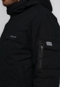 Schwarze isolierte Jacke mit einer Reißverschlusstasche am linken Arm und einem etikettierten Aufnäher. Verfügt über eine Kapuze und eine glatte Stoffoberfläche.