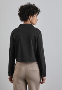 Kort zwart jasje van een gestructureerde stof, met een klassieke kraag en een knoopsluiting bij de manchet, in combinatie met beige broeken en achterzakken.