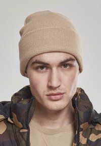 Beige strikket hue med ribbet struktur, båret slouchy. Model i en camouflagejakke og beige sweater. Neutral baggrund.