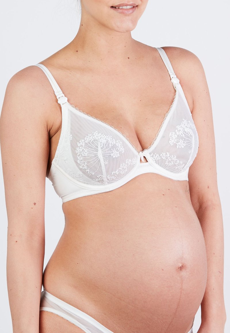 Pregnant woman wearing white lace bra and matching underwear, showing her rounded belly and a subtle smile against a plain background.