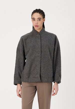 Giacca in fleece grigia con colletto alto, chiusura frontale con bottoni e maniche ampie. Tessuto morbido con una vestibilità rilassata, abbinata a pantaloni marroni.