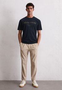 Marineblaues Baumwoll-T-Shirt mit goldenen Aufdruck, kombiniert mit beigefarbenen Hosen und weißen Sneakern. Das Model steht vor einem neutralen Hintergrund.