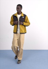 Giacca in pile con pannelli gialli e neri, tasche con zip, abbinata a pantaloni cargo beige. In piedi su un pavimento blu chiaro.