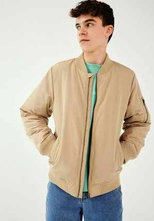 Beige bomberjacka med ribbad manschetter och nederkant, med dragkedja och sidofickor, bärs över en teal-färgad skjorta.