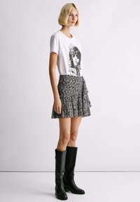 Weißes T-Shirt mit schwarzem Grafikdruck, kombiniert mit einem gemusterten, rüschierten Minirock in dunklen Farben. Knielange schwarze Lederstiefel.