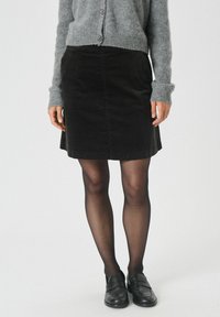 Zwarte corduroy rok met een aansluitende taille en een lichte A-lijn vorm. Gepaarde met een grijze trui en zwarte schoenen. Transparante panty gedragen.