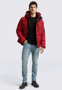 Rød dunjakke med hætte, sorte detaljer og lynlåslommer, kombineret med lyseblå jeans og sorte ankelstøvler. Glat tekstur over det hele.