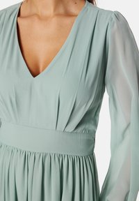 Mintgroene jurk met een diepe V-hals, lange doorzichtige mouwen, geplooid lijfje en een aansluitende tailleband die leidt naar een vloeiende rok.