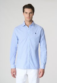 Camicia a maniche lunghe di colore azzurro chiaro con strisce verticali bianche, dotata di un piccolo logo ricamato sul petto e colletto a bottoni.