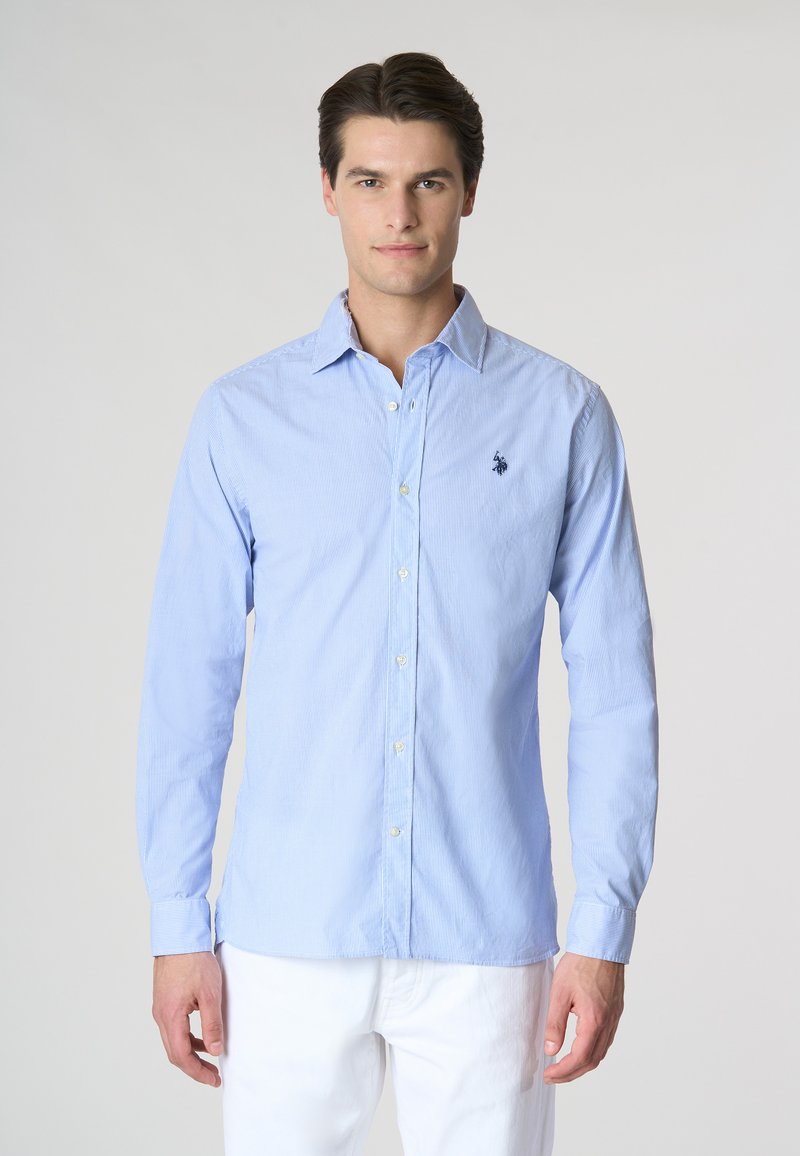 Camicia a maniche lunghe di colore azzurro chiaro con strisce verticali bianche, dotata di un piccolo logo ricamato sul petto e colletto a bottoni.
