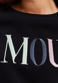 Svart sweatshirt med texturerad broderi med ordet "MOUT" i ljusgrön, mörkblå och rosa, mot en slät tygbakgrund.