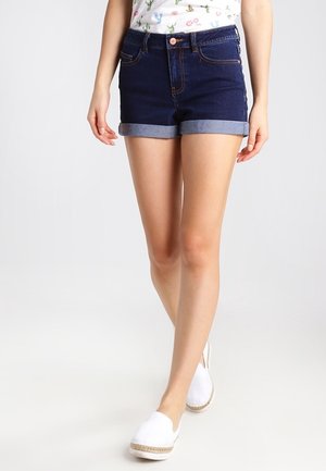 Persoon die donkerblauwe omgeslagen spijkershorts, een wit top met print en witte instapschoenen draagt, lopend vooruit op een effen achtergrond.