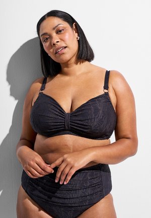 Frau mit glatten schwarzen Haaren, die einen schwarzen BH mit floraler Struktur und passende hoch taillierte Unterwäsche trägt, steht vor einer weißen Wand.