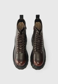 Egy pár sötétbarna bőr fűzős csizma vastag fekete talppal és oldalsó cipzárral, felülről nézve, fehér háttér előtt.