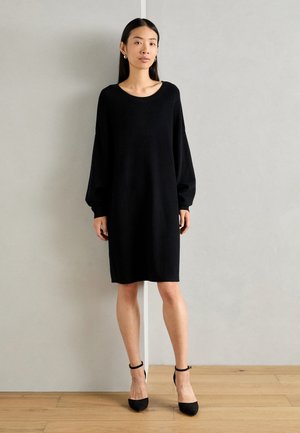 Abito nero a maglia con vestibilità comoda, scollo rotondo e maniche lunghe a palloncino. Indossato con tacchi neri e posizionato su uno sfondo neutro.