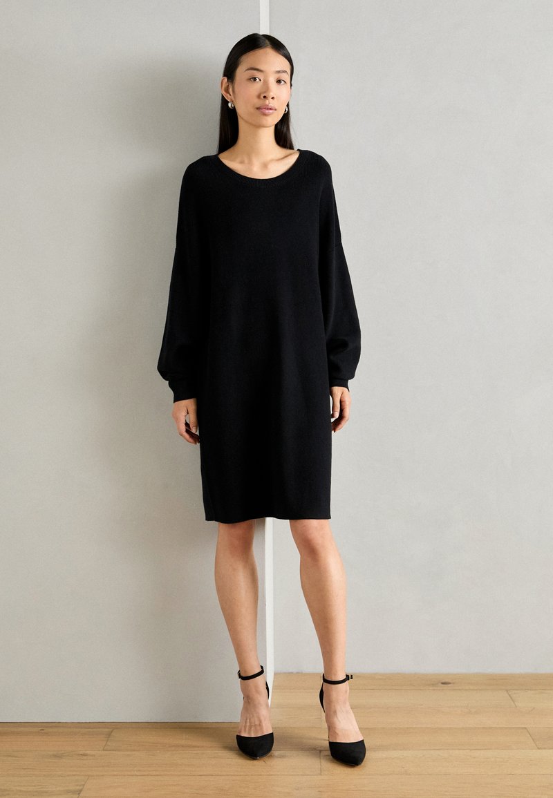 Black knit dress with a relaxed fit, round neckline, and long, balloon-style sleeves. Worn with black heels and set against a neutral background.