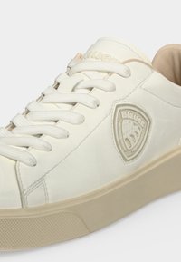 Baskets basses blanches avec semelle beige, lacets blancs et écusson du logo Blauer sur le côté. Aucun détail d’arrière-plan visible.