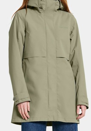 Kobieta w jasie przeciwdeszczowym w jasnym oliwkowo-zielonym kolorze z kapturem i zapinanymi na guziki mankietami, w zestawieniu z niebieskimi dżinsami na jednolitym tle.