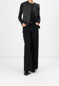 Černá prošívaná vesta přes černé tričko s dlouhým rukávem, spárovaná s černými kalhotami s vysokým pasem a širokými nohavicemi a černými botami s texturovaným detailem.