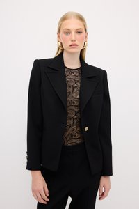Blazer nero con un design strutturato, caratterizzato da un collo a risvolto e un singolo bottone dorato. Indossato su una maglietta in pizzo a fantasia.