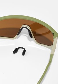 Sport-Sonnenbrille mit einem grün-creamfarbenen Rahmen, braunen Gläsern und schwarzen Gummi-Nasenpads. Inklusive abnehmbarem schwarzen Riemen.