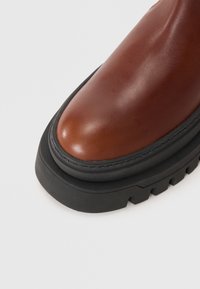 Stivale ankle in pelle marrone con punta rotonda, dotato di una spessa suola in gomma nera e cuciture a contrasto lungo il bordo.
