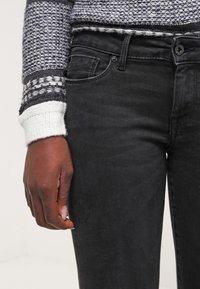 Jeans de denim negro con un corte slim, que presentan un diseño clásico de cinco bolsillos, y un suéter gris con un patrón y un tejido textura, además de detalles en los puños.