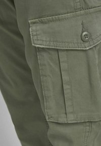 Gros plan sur la poche d’un pantalon cargo vert olive avec un rabat fermé par un bouton à quatre trous et des détails de couture visibles.