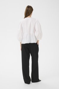 Hvid bluse med puffærmer, bølget kantdetalje, bomuldsmateriale og rund halsudskæring, kombineret med løse sorte bukser.