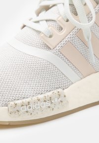 Baskets tricotées gris clair avec des accents beiges, présentant une tige texturée, des rayures blanches et une semelle mouchetée avec un logo en relief.