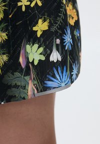 Nærbilde av et svart tøy skjørt med fargerike blomsterdesign i blått, gult, hvitt og grønt, båret over et bare bein.