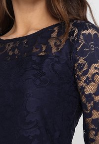 Námořnické modré krajkové šaty s květinovými vzory a průhlednými dlouhými rukávy, s pevným korzetem a texturovanou tkaninou pro větší hloubku.