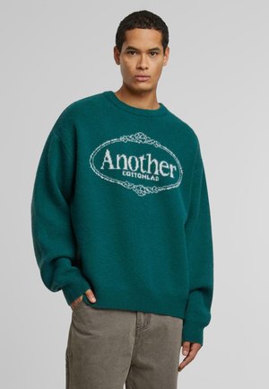 Suéter de algodón verde con corte oversize, cuello redondo y logotipo blanco "Another Cottonlab" enmarcado en un óvalo. Puños y dobladillo acanalados.