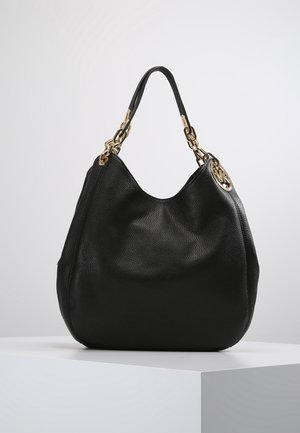 Sac à main en cuir noir avec des maillons de chaîne dorés et un pendentif rond doré avec logo, présenté sur un socle blanc sur un fond gris clair.