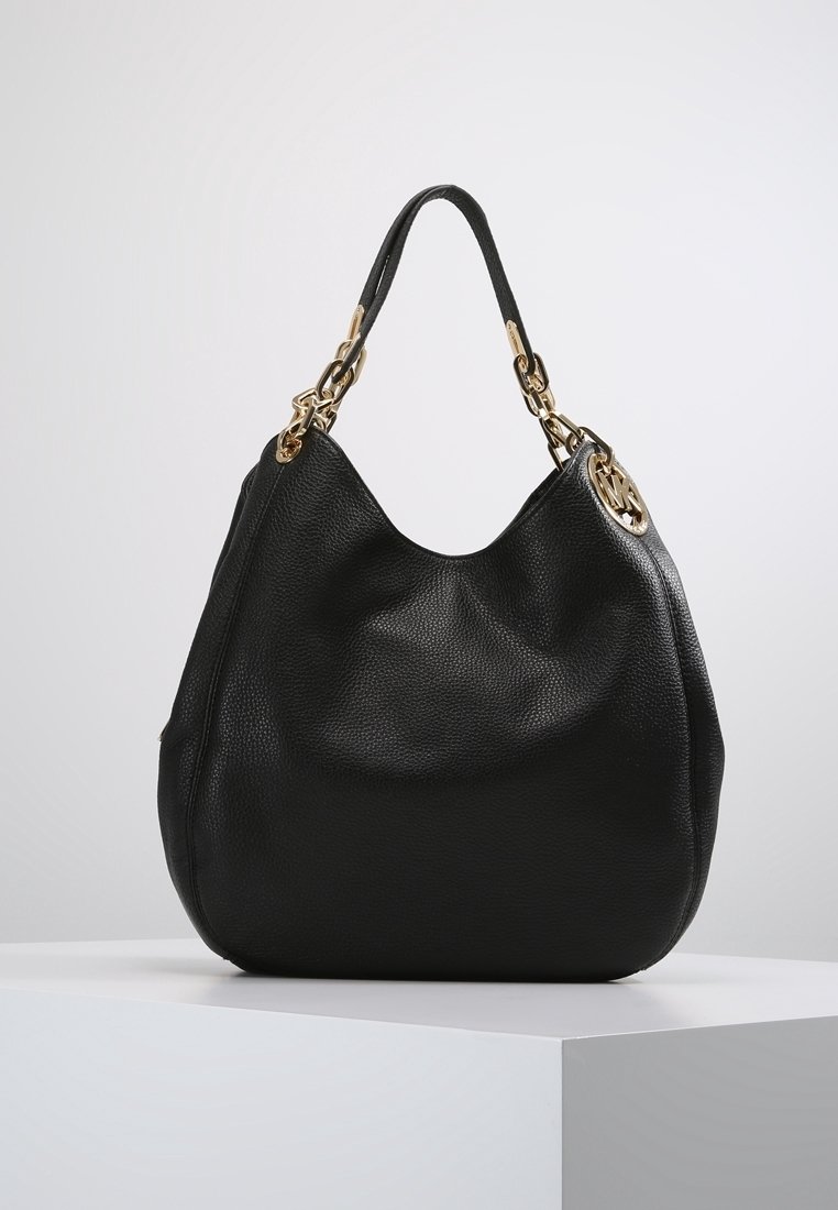 Sac à main en cuir noir avec des maillons de chaîne dorés et un pendentif rond doré avec logo, présenté sur un socle blanc sur un fond gris clair.