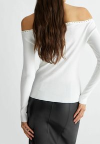 Pull blanc à épaules dénudées en côtes, à manches longues, orné de clous décoratifs le long de l'encolure, associé à un pantalon noir ajusté.