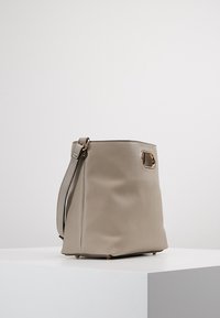 Vaalean beige nahkainen ämpärilaukku, jossa on sileä pinta. Laukussa on tukeva hihna, kultaiset yksityiskohdat ja litteä pohja.