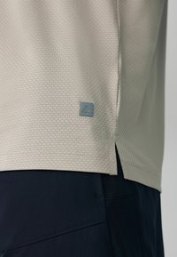 Ourlet de chemise texturé beige avec un petit logo triangulaire gris au-dessus de la fente latérale, porté sur un pantalon bleu marine foncé.