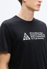 Μαύρο βαμβακερό μπλουζάκι με λευκό γραφικό που απεικονίζει ένα τρίγωνο και κείμενο λεπτομέρειας. Λαιμόκοψη crew και κοντά μανίκια.