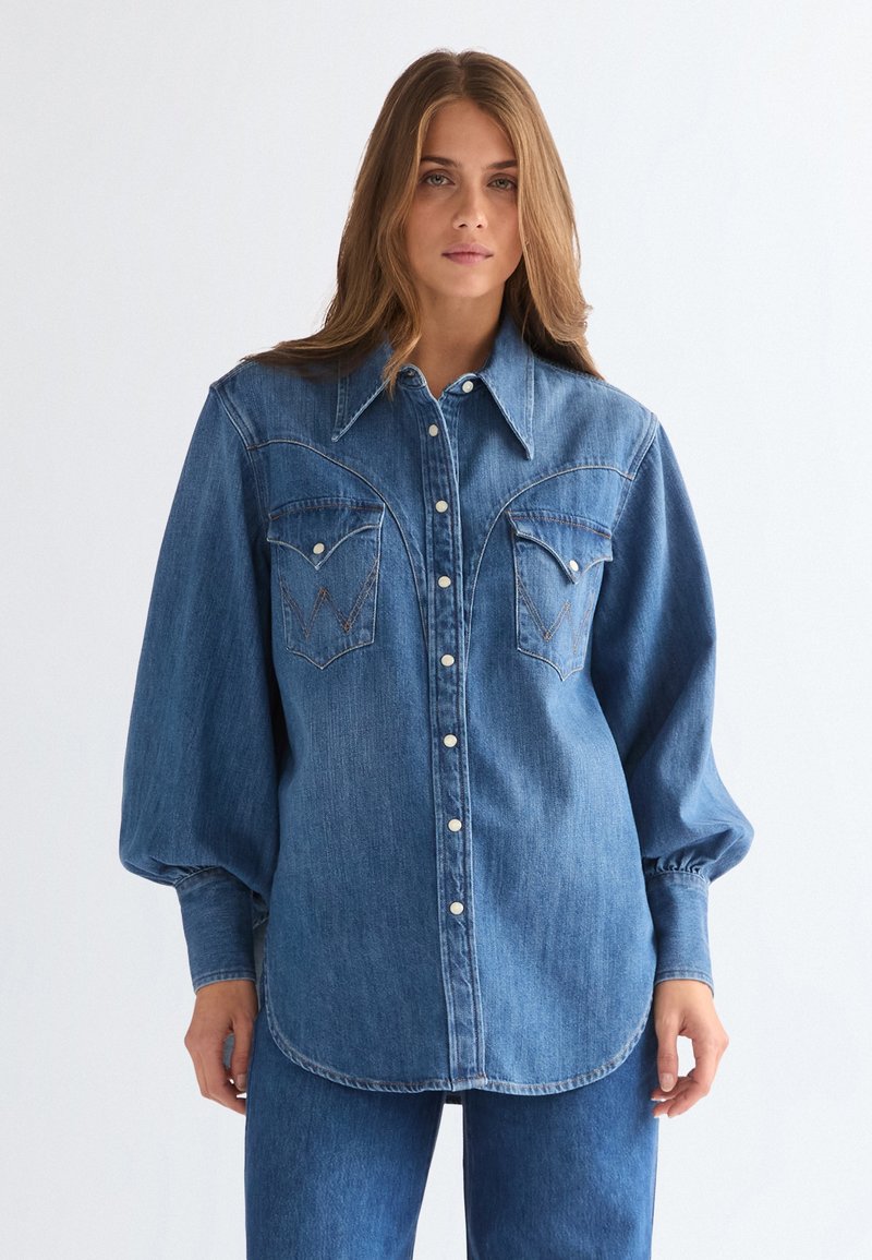 Νεαρή γυναίκα με μακριά μαλλιά, ντυμένη με μπλε τζιν πουκάμισο με φουσκωτά μανίκια, κουμπιά κλιπ και ταιριαστό τζιν παντελόνι, σε λευκό φόντο.