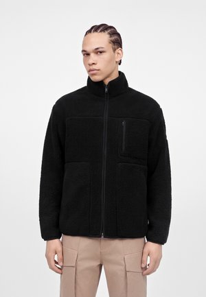 Joven con el pelo trenzado que lleva una chaqueta polar negra con cremallera y pantalones beige sobre un fondo blanco liso.