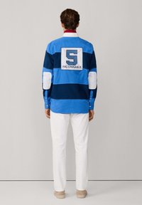 Blauwe en marine gestreepte langeblouse met witte accenten en een geborduurd logo, gecombineerd met witte broek en beige schoenen.