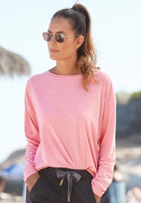 Helles rosa Langarmshirt mit lockerer Passform, Rundhalsausschnitt und überschnittenen Schultern, kombiniert mit schwarzen Jogginghosen mit Kordelzug und Sonnenbrille.