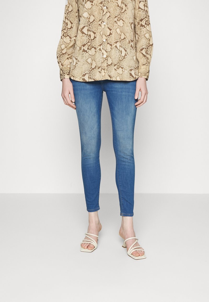 Blusa beige con estampado de serpiente y mangas largas, combinada con jeans ajustados azules y sandalias beige con tiras. El conjunto tiene un corte relajado.