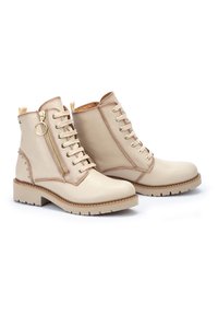 Botas de tobillo de cuero beige con un acabado texturizado, detalles en dorado y cordones. Cuentan con una cremallera lateral y tachuelas en el tacón. Suela plana.