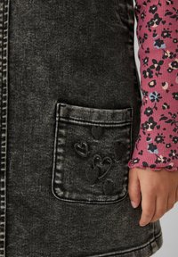 Zwarte denim jurk met een voorkleurzak met geborduurde hartontwerpen. Een roze bloes met lange mouwen is gedeeltelijk zichtbaar bij de mouw.