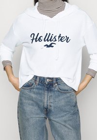 Sweat à capuche blanc en tissu texturé affichant "Hollister" en lettres bleu marine, associé à un jean en denim bleu clair. Col roulé rayé en-dessous.