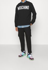 Sweat-shirt noir avec le logo "MOSCHINO", superposé à une chemise en denim bleu clair. Pantalon cargo noir et baskets multicolores à semelles épaisses.