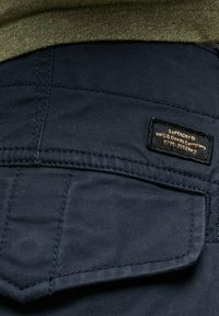 Tessuto blu scuro dei pantaloni con una finitura testurizzata. Presenta una tasca posteriore e un'etichetta rettangolare del marchio cucita sulla tasca.